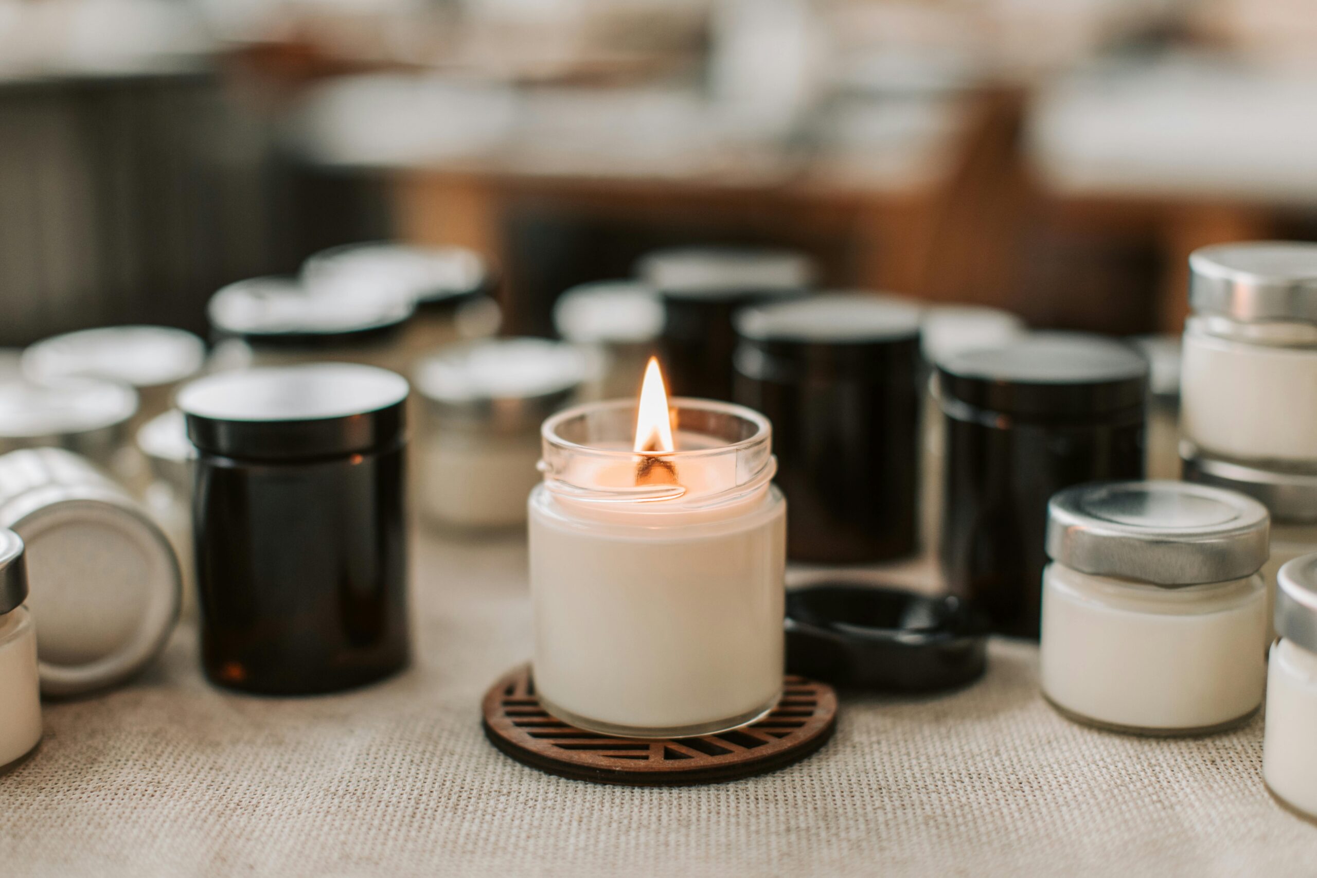 A close-up of a handmade white candle with a burning flame, surrounded by candle jars on a table.