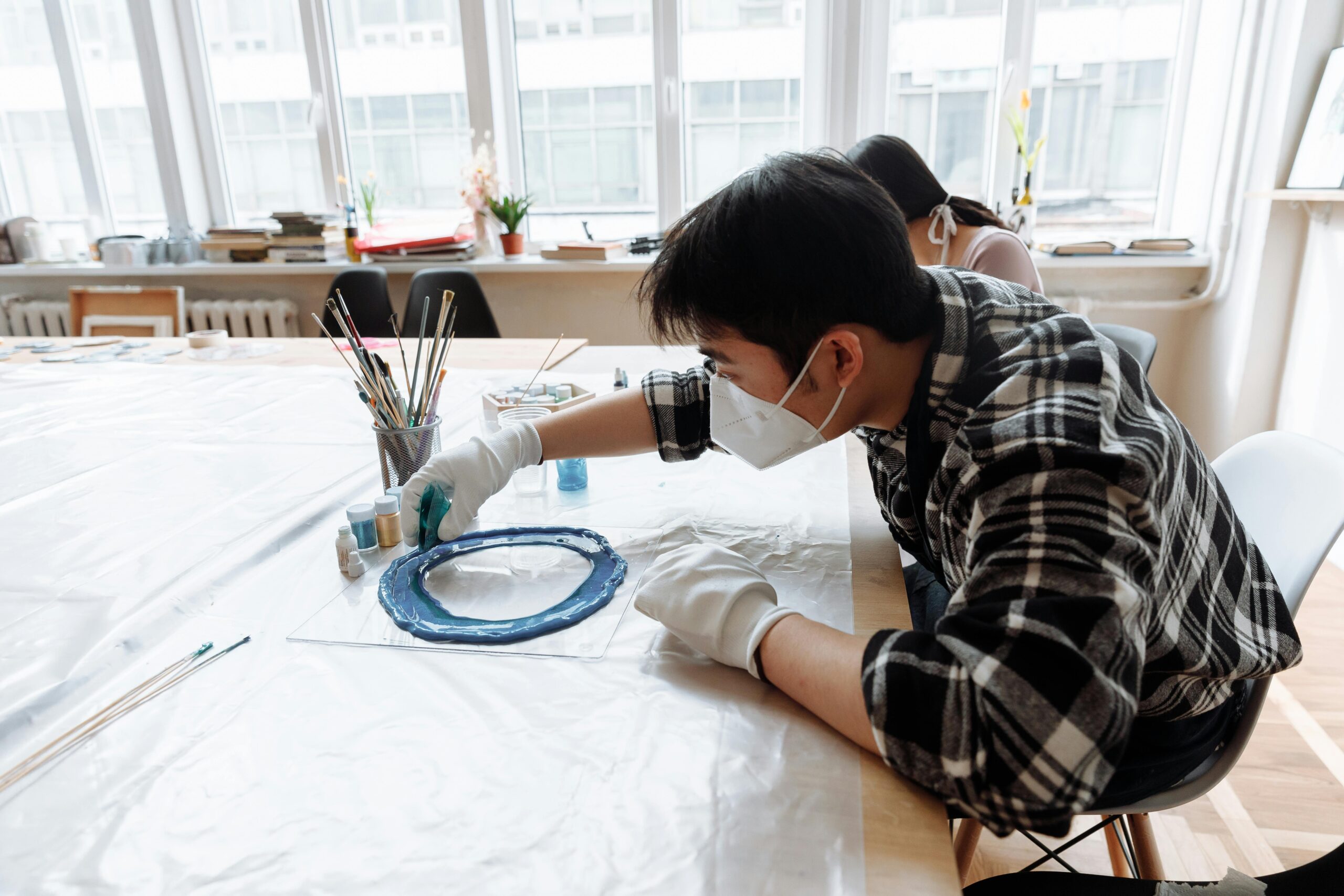 Asian man wearing face mask creating resin art at a bright indoor workspace.