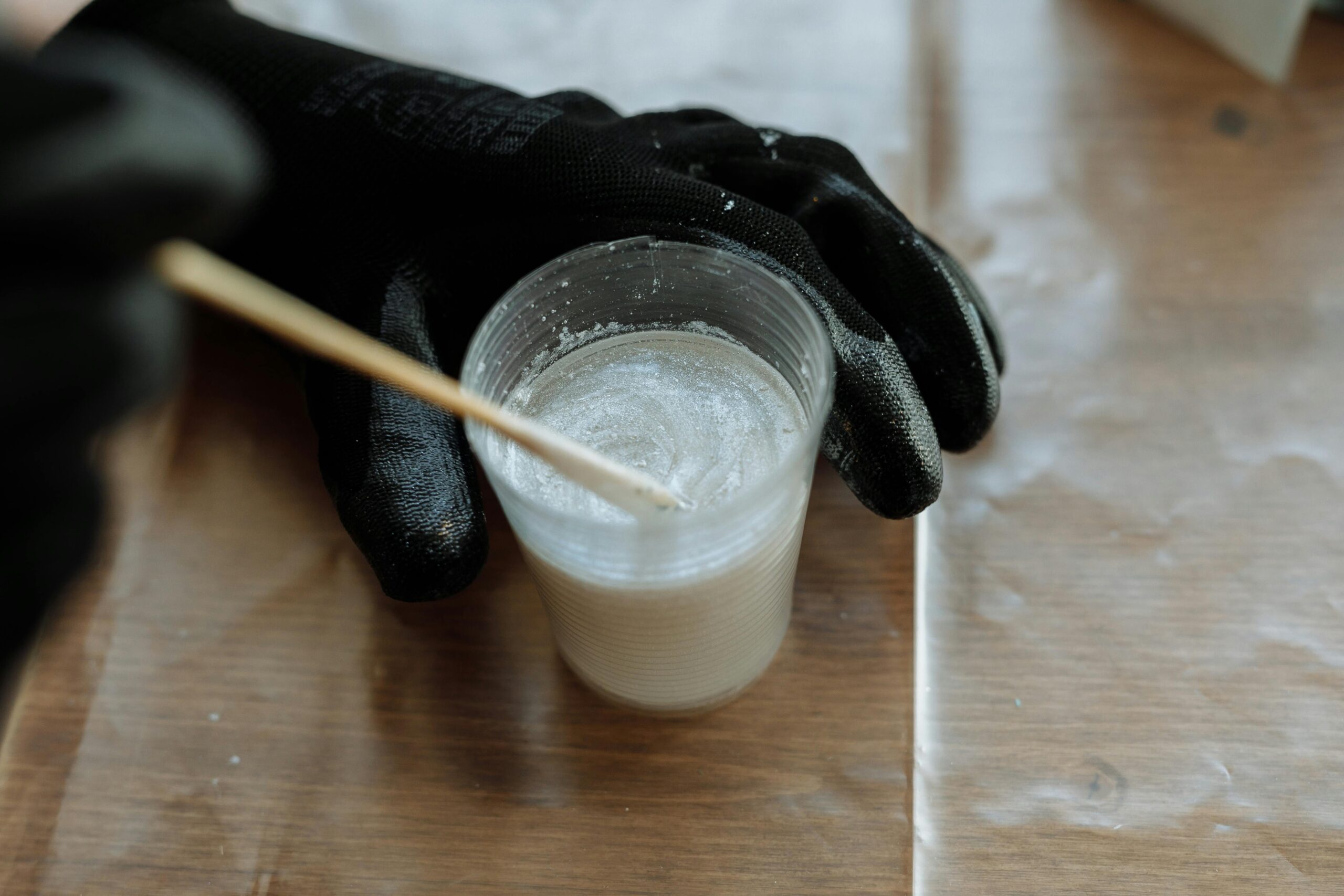 Detailed view of hands with gloves mixing resin in a plastic cup with a stirrer on a wooden surface.